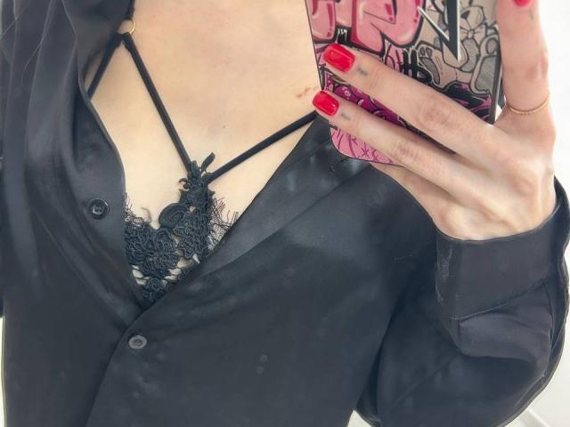 Close-up of MoonLiza's chest, wearing a black button-up shirt and lace bralette, hand with red nails holding phone with manga design, visible cleavage, black straps, skin tone is pale.