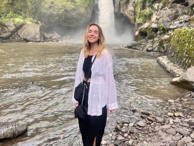 RoxyFoxy-hot está de pie frente a una cascada, sonriendo, con el cabello rubio ondulado y mojado. Ella lleva una blusa blanca, pantalones negros y lleva un bolso negro. Increíblemente atractiva en un entorno natural..