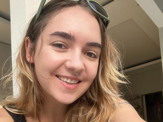 Fotografía de primer plano de RoxyFoxy, atractiva, con cabello rubio ondulado, piel clara y un piercing en la nariz. Ella está sonriendo, lleva gafas de sol en la cabeza y tiene las cejas marrones. Vibración casual, sexy y segura..