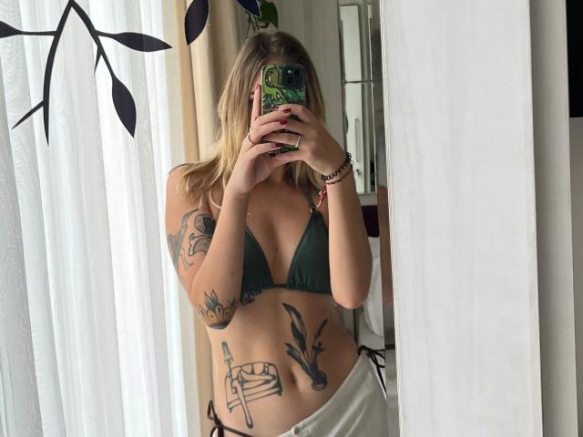 RoxyFoxy: bellissima, con lunghi capelli biondi, un top da bikini nero e pantaloni bianchi, tiene in mano un telefono che le copre il viso, con tatuaggi sul torso e braccia, luce solare intensa, finestra con una pianta verde sullo sfondo. Molto attraente..