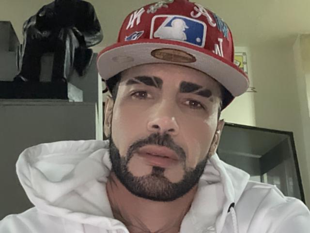 Autoritratto ravvicinato di DarwinHot con un berretto da baseball rosso, una felpa bianca e una barba folta. L'espressione seria di DarwinHot e le sue labbra leggermente dischiuse suggeriscono tensione. Giacca nera sullo sfondo.