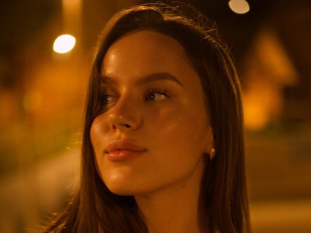 Close-up de MichaMattes, uma jovem com cabelos longos e escuros, pele brilhante e lábios cheios, olhando para o lado com um expressão sedutora, banhada em uma luz quente e alaranjada. Fundo desfocado.