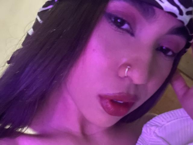 Gros plan sur Angels69v, une jeune femme à la peau légèrement violette, aux cheveux foncés et portant un anneau au nez, qui regarde de manière séduisante. caméra. Elle porte un bandeau blanc et un haut blanc, avec du rouge à lèvres..