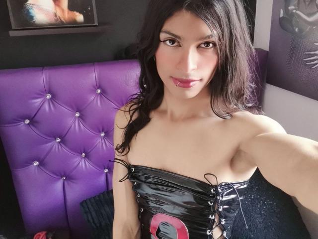 LunarisSmith, une femme aux longs cheveux noirs, vêtue d'une robe en latex noire sans bretelles, est assise contre un dossier violet capitonné. Tête de lit. Elle sourit avec un regard sexy et confiant. Ses seins sont petits..