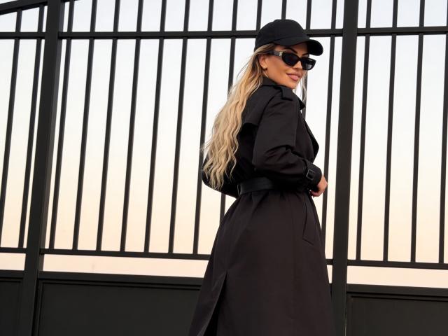 ZoeyMarie staat met haar gezicht naar links, ze draagt een zwarte muts en zonnebril, haar lange blonde haar wappert in de wind, een zwart trenchcoat, zwart hoed, kijkt over de schouder met een zelfverzekerde, aantrekkelijke uitdrukking. Balken op de achtergrond.