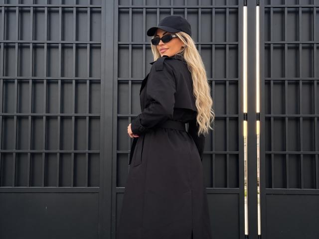 ZoeyMarie, com cabelos loiros longos, boné preto e óculos de sol, vestindo um casaco preto longo, está em pé em frente a um padrão quadriculado. parede preta.
ela olha por cima do ombro, transmitindo um charme confiante..