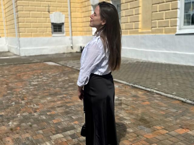 MichaMattes se tient sur une rue pavée, vue de profil, portant une chemise blanche et une jupe noire, avec de longs cheveux bruns. Écartant le regard.
fond d'un bâtiment jaune et blanc, ensoleillé, avec des pavés ombragés. Extrêmement sexy..