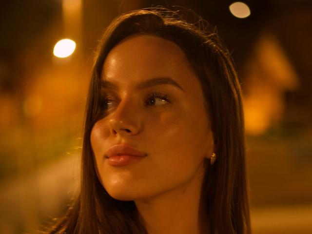 Close-up photo of MichaMattes, a dark-haired woman with smooth skin and full lips, illuminated by warm, yellow streetlights. Her gaze is slightly up, with a hint of sexual tension.