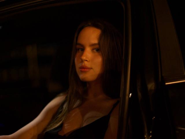 Photograph of MichaMattes with long brown hair, sitting in a car at night, looking out with a serious expression. Dim lighting casts shadows on her face, highlighting her focused gaze.