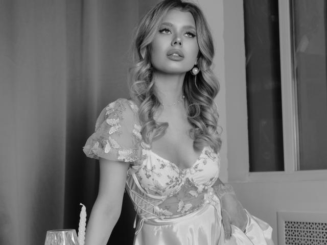 Fotografía en blanco y negro de FollowLove con cabello largo y ondulado, vestida con un vestido de corte bajo con encaje, de pie contra una ventana. FollowLove tiene una expresión segura y sensual, con escote visible..
