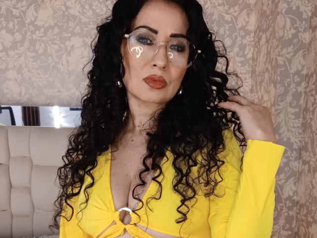 Samantha Antolin, com longos cabelos cacheados e pretos, vestindo uma blusa amarela com botões e decote profundo, e um colar em formato de coração. adesivos em óculos, e batom vermelho vibrante. O fundo tem um papel de parede com padrão floral. Incrivelmente sexy..