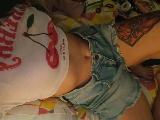 Isabella Palacio está acostada en una cama, vistiendo una camiseta blanca de "Coca-Cola" y pantalones cortos vaqueros desgastados. Su abdomen está al descubierto, mostrando un ligero bronceado. Tiene un tatuaje en la parte interna de su muslo derecho. Increíblemente atractivo/a..