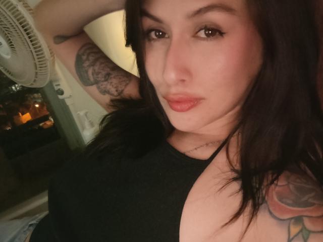 Selfie de perto de IsabellaPalacio, de pele clara, com cabelo escuro, olhos castanhos e lábios vermelhos, vestindo uma blusa preta, com a mão atrás da cabeça, exibindo uma tatuagem detalhada no braço superior, tensão sexual.