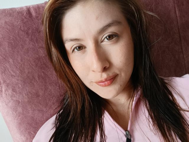 Primer plano de Isabella Palacio, una joven con cabello negro lacio, piel clara y ojos marrones, sonriendo ligeramente. Ella lleva una camisa rosa y está acostada sobre un cojín morado. Increíblemente sexy..