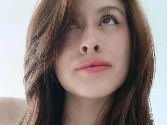 Close-up de Isabella Palacio, de pele clara, com cabelo castanho cobrindo um olho, olhos azuis olhando ligeiramente para o lado. lábios rosados, expressão suave, bochechas levemente coradas, maquiagem mínima, fundo neutro..