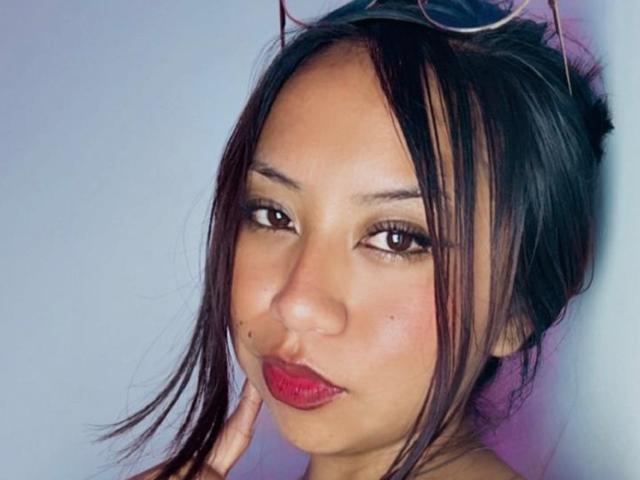 Close-up de Emmilyy69 com cabelo preto desgrenhado, batom vermelho e uma pequena marca de beleza na bochecha. Ela olha de forma sedutora, os dedos tocando seu rosto, com seus intensos olhos castanhos. Muito quente.