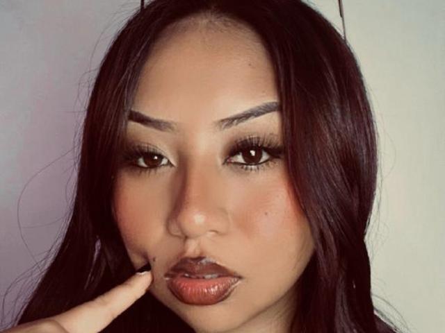 Close-up de Emmilyy69, uma mulher asiática com cabelos longos e pretos, sobrancelhas grossas e batom vermelho escuro, tocando o queixo. com o dedo indicador. Seus olhos estão fortemente maquiados, com uma expressão sedutora..