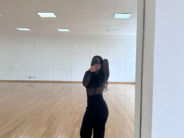 IanaPrince prend un selfie devant un miroir dans une pièce lumineuse et vide, avec des planchers en bois. Elle porte un haut noir transparent et un pantalon noir, et elle cache son visage avec un téléphone. Ses longs cheveux noirs sont attachés en une queue de cheval.
.