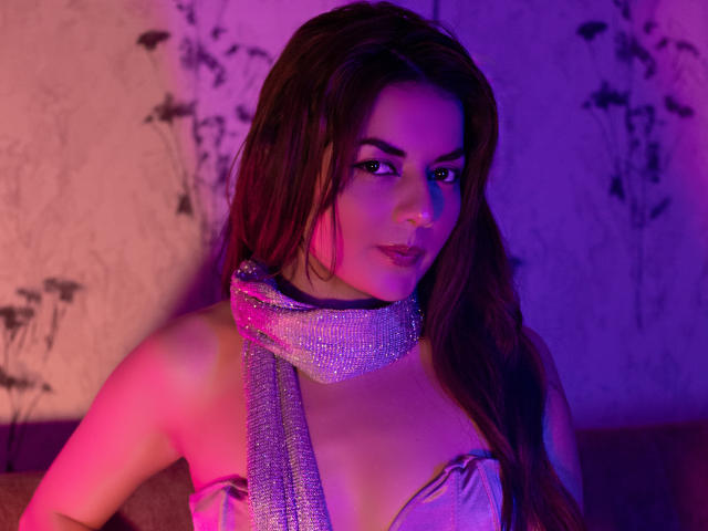 Las MandyMouses, con cabello largo y castaño, que llevan una bufanda plateada brillante y una blusa con escote pronunciado, están iluminadas con colores púrpura y rosa. iluminación. Mirada seductora, ligera sonrisa. El fondo muestra patrones florales desenfocados..