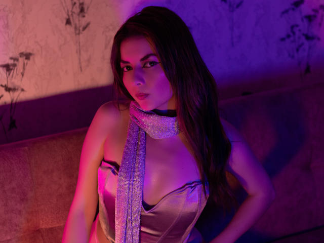 MandyMouses con lunghi capelli castani, che indossano un abito rosa scintillante e una sciarpa luccicante, sedute su un divano sotto un [purple and] illuminazione al neon rosa, che guarda direttamente nella fotocamera con un'espressione seducente.