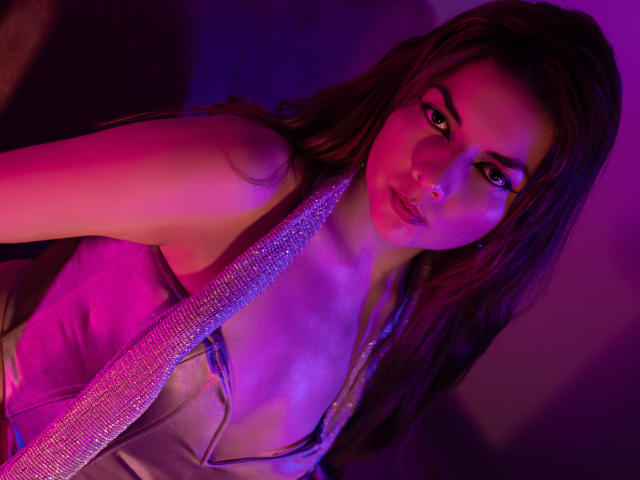 Fotografía de MandyMouses, con cabello largo y castaño, vistiendo una camiseta de tirantes con escote pronunciado, iluminada por luces rosas y azules. Ella sostiene una corbata rosa brillante, mirando directamente a la cámara con una expresión seductora..