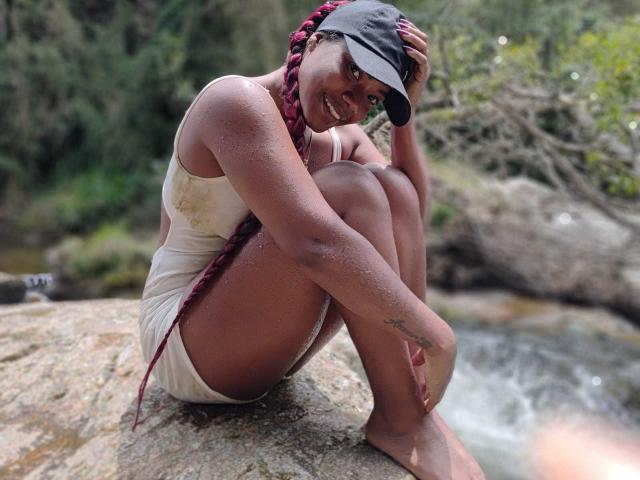Caylin está sentada en una roca, con las piernas dobladas, vistiendo una camiseta blanca mojada y una gorra negra, y sonriendo. Trenzas que caen sobre los hombros, gotas de agua en la piel, fondo de bosque. Increíblemente atractivo/a..