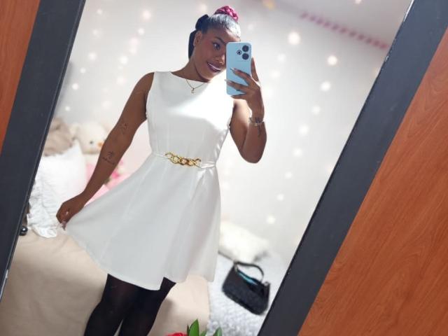 Caylin está em frente a um espelho, segurando um smartphone, vestindo um vestido branco sem mangas com um cinto dourado, preto calças legging e uma tiara rosa com uma flor. Fundo claro e refletor. Sorrindo.