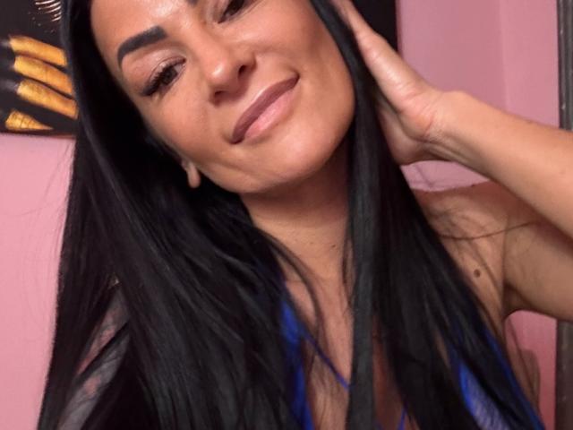 Close-up of AndraD, a woman with long black hair, smiling slightly, hand touching her face, pinkish skin, subtle blue highlights in hair, confident expression, casual selfie vibe, sexual tension present.