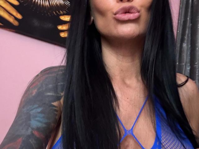 Gros plan d'AndraD avec de longs cheveux noirs, des lèvres pulpeuses, portant un haut en maille bleu avec des détails à bretelles fines, mettant en valeur un tatouage. épaule. Sa poitrine est légèrement dénudée, ce qui crée une ambiance séduisante..