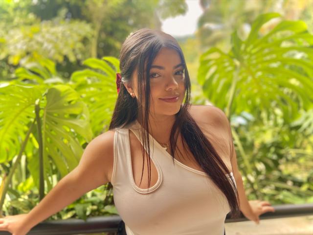 Lara James, con su largo cabello negro, se apoya en una barandilla, vistiendo una camiseta blanca y mojada que se adhiere a su cuerpo. senos, rodeados de un follaje verde brillante. La luz del sol resalta su piel húmeda.