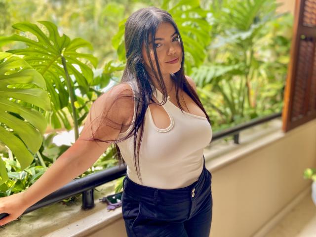 Lara James está em um ambiente externo exuberante e verde, apoiada em um corrimão preto. Ela tem cabelos longos e pretos, veste uma camiseta branca justa e calças pretas de cintura alta. A luz forte do sol realça suas curvas e seus seios grandes..