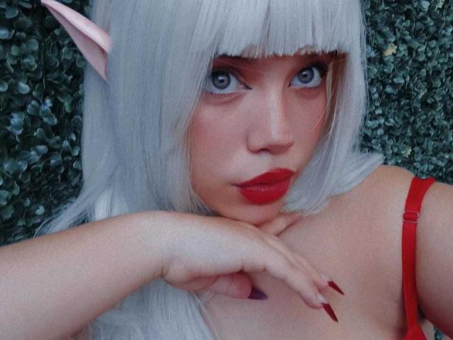 Fotografía de primer plano de AryButty con orejas de elfo blancas, piel pálida y lápiz labial rojo. Ella la mira de forma seductora con sus ojos azules, apoyando su mano en su pecho, y lleva una correa roja..