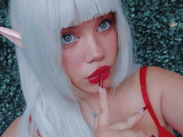 Foto em close-up de AryButty com cabelo branco pálido, orelhas de elfo e batom vermelho. Ela está mordendo o dedo, com os olhos azuis arregalados, vestindo uma blusa vermelha com alças. Olhar incrivelmente atraente e intenso..