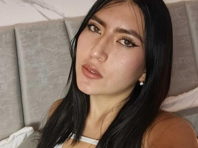 Fotografía de primer plano de EmmaRouxx, una mujer atractiva con cabello negro largo, ojos marrones y labios carnosos. Ella lleva una blusa blanca y mira a la cámara de forma seductora. Fondo de cabecera tapizada en color gris..