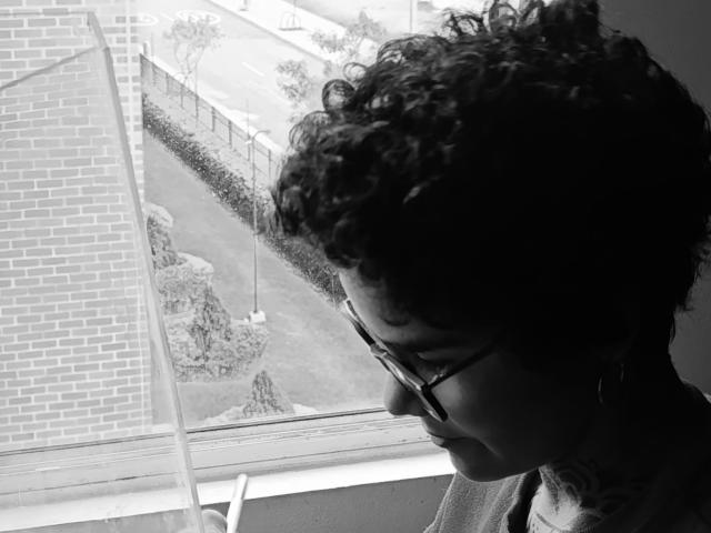 Black and white photo of AnaRoa with curly hair and glasses, looking out a window. AnaRoa's face is shadowed, cocked head, intense gaze. Bright window and brick wall in background.