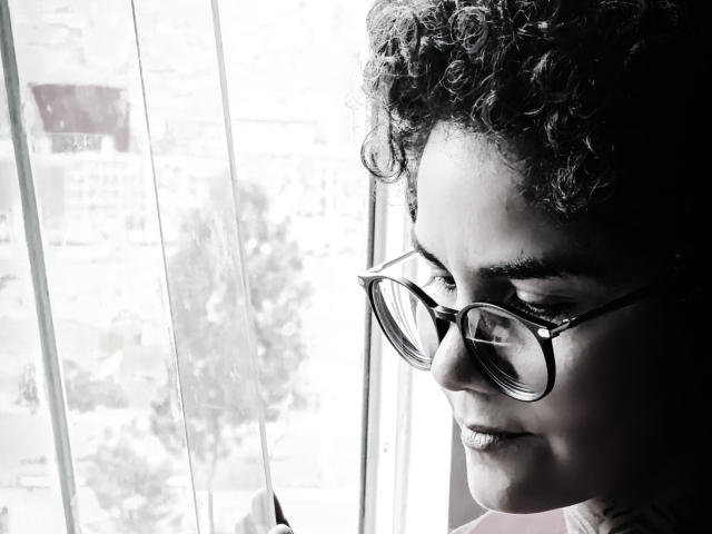 Close-up photograph of AnaRoa, a curly-haired person with glasses, looking down with a slightly melancholic expression. Bright light from the window contrasts with dark shadows on their face. High contrast.