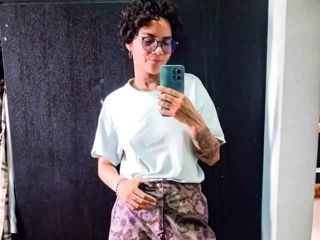 AnaRoa, kort krullend zwart haar, bril, wit T-shirt, houdt een groene telefoon vast, bloemenbroek, staat voor een zwarte achtergrond. behandelde muur, subtiele glimlach, rechterhand op de heup, nonchalante spiegelselfie, moderne stijl.