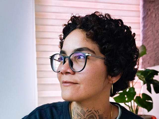 Photograph of AnaRoa, a curly-haired person with glasses, looking to the side. Tattoo on neck, black shirt, pinkish sunlight background. Focused expression, slight smile, visible ear piercing.