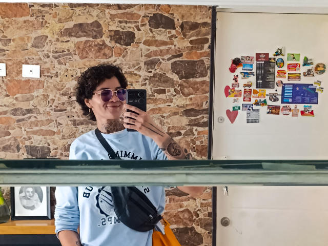 AnaRoa, de cabelos cacheados, usando óculos de sol e uma camiseta branca com uma bolsa transversal preta, tira uma selfie em frente a parede de tijolos, geladeira com ímãs coloridos ao fundo. Muito atraente..