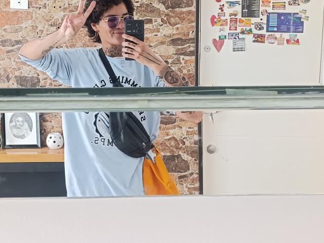 Photograph of AnaRoa, standing in front of a mirror, making a peace sign with her right hand, wearing a white T-shirt, black crossbody bag, orange bag, and dark sunglasses.