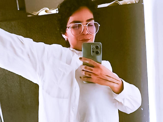 AnaRoa maakt een selfie in een auto, gekleed in een wit shirt en met grote, ronde brillen. Een telefoon vasthouden met een zichtbare camera, arm uitgestrekt, lichte grijns. Hel licht accentueert het gezicht.