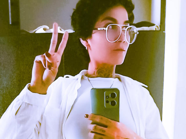 AnaRoa, short dark hair, wearing oversized white glasses, white shirt, holding a smartphone, flashing a peace sign with left hand, bright light behind, slightly blurred, casual selfie, modern style.