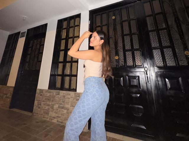 SarahLanne, una donna formosa con lunghi capelli castani, indossa un top corto bianco e pantaloni di pizzo azzurri, è in piedi di fronte a porte in vetro nero, una mano che tocca il suo viso, posa sensuale.
