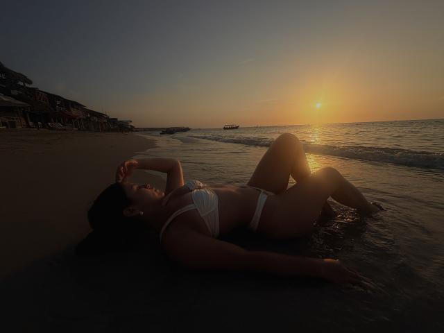 SarahLanne está tumbada en una playa al atardecer, vestida con un bikini blanco. Está boca arriba, con una mano en la cabeza, las piernas dobladas, silueteada contra el sol y el océano brillantes..