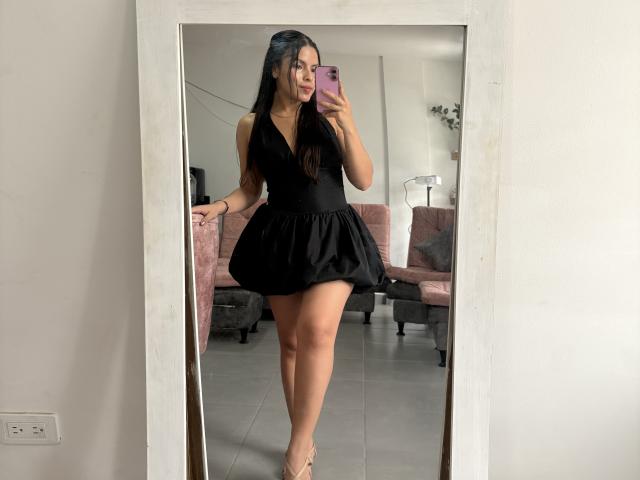 SarahLanne, con i suoi lunghi capelli neri, indossa un abito corto nero con un profondo scollo a V e si scatta un selfie allo specchio. Sorride, mostrando un accenno di scollatura, e tiene in mano un telefono rosa. Molto attraente..