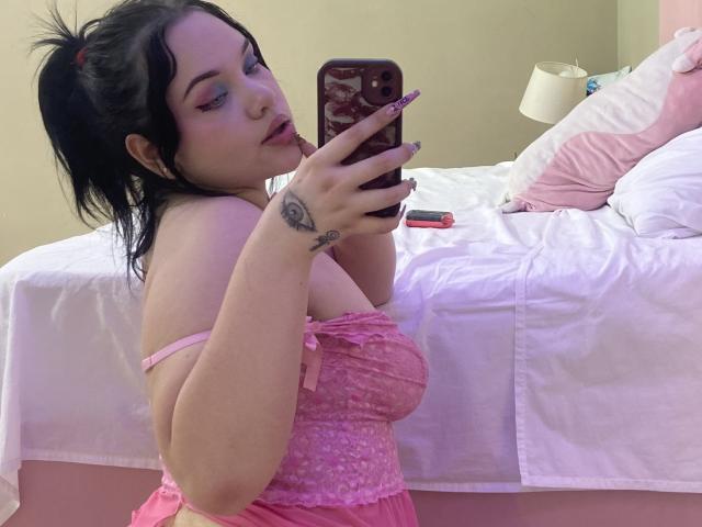 AlayaWillow mit schwarzen Haaren im Pferdeschwanz, trägt eine rosa Spitzenbluse und macht ein Selfie im Spiegel. Sie hat Tattoos an ihrem Handgelenk und leckt andeutungsweise ihre Lippen. Bett mit weißen Laken im Hintergrund.