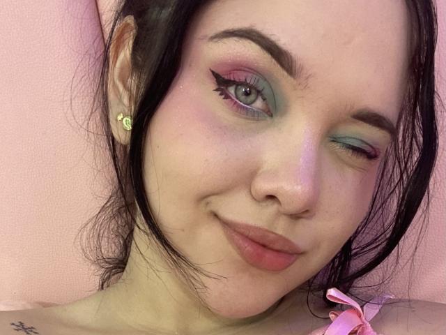 Close-up of AlayaWillow winking, pale skin, black hair, pink and green eyeshadow, black eyeliner, red lipstick, small yellow earrings, pink flower in hair, slight smirk, soft pink background, visible tattoo on neck.