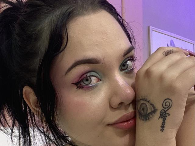 Fotografia ravvicinata di AlayaWillow, con la pelle chiara, capelli neri, ombretto blu e rossetto rosa. Lei si sta mordendo il dito, rivelando un tatuaggio con la scritta "Soul" sulla mano destra. Uno sguardo incredibilmente attraente e intenso..