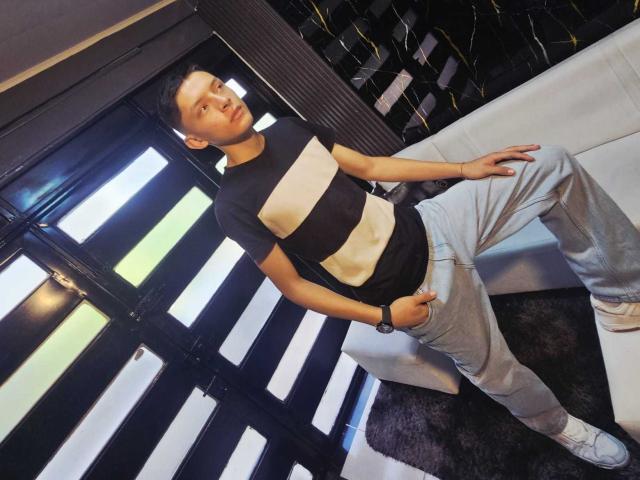 SamuJasper, standing casually, leaning against a white couch, wearing a black and white striped shirt and light gray pants, white sneakers, dark hair, black wristband, bright window light, black floor.