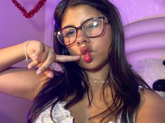 Foto em close-up de CelesteMorel, com cabelo escuro, usando óculos e uma blusa branca, com a língua para fora enquanto toca. Ela tocou com o dedo. Ela tem uma pulseira e um colar de prata. Incrivelmente sexy..